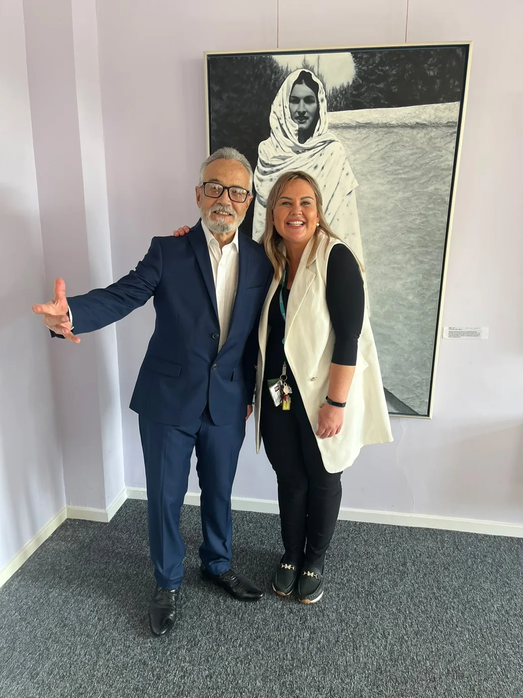 Max Nadimi in a navy suit, one arm stretched out, standing beside Kasia Luczynska in front of a large black and white painting of a woman in a white shawl.